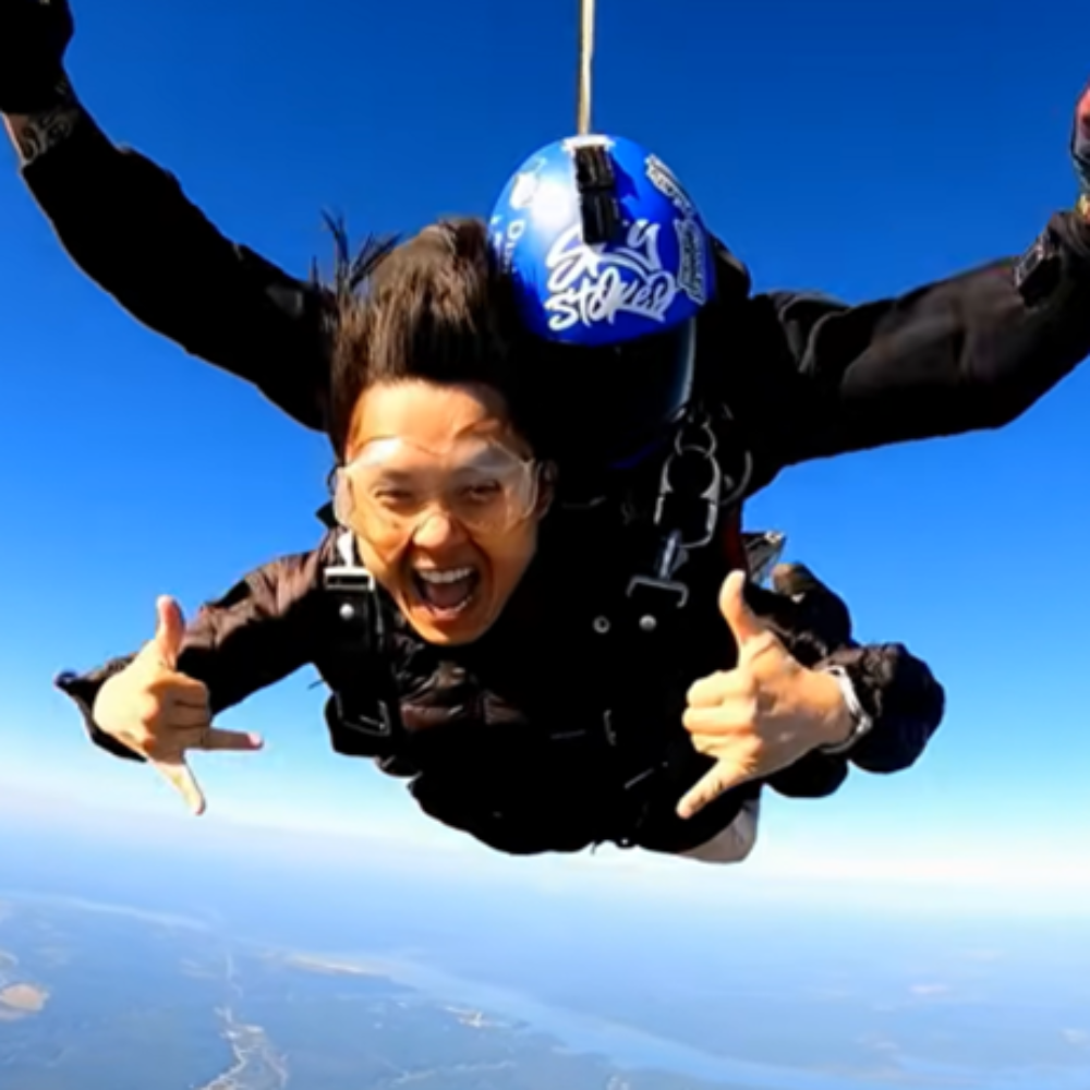 Dr. Tran skydives in tandem with an instructor, smiling widely and making shaka hand signs (pinky and thumb extended)