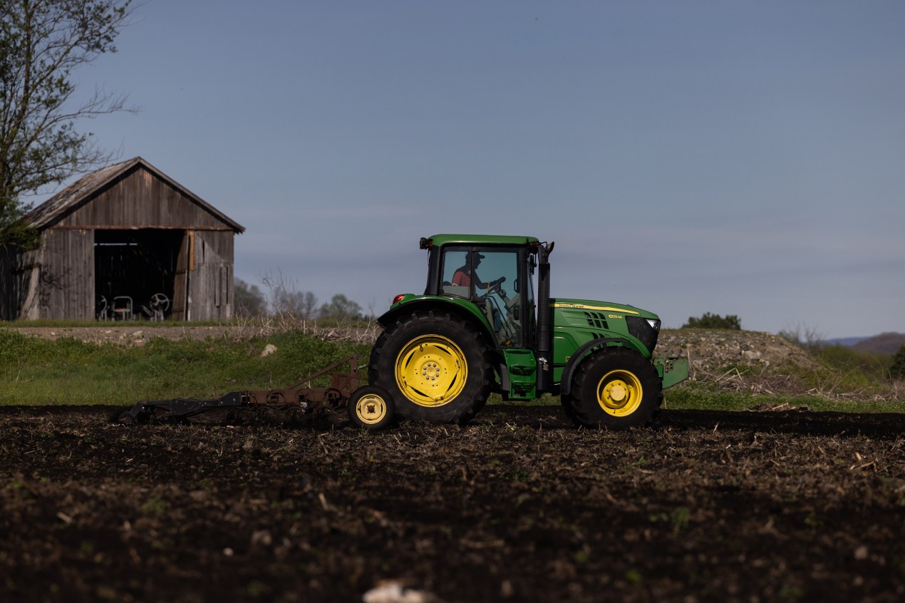 A barn is seen behind a man driving a green tractor across a field