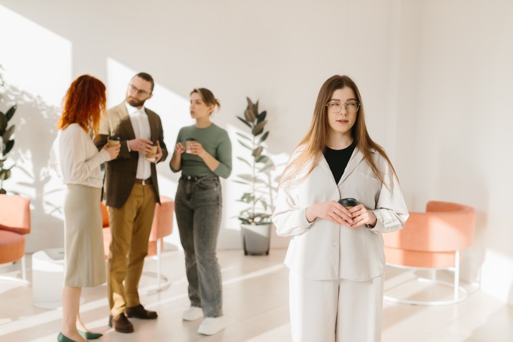 A woman rolling her eyes while holding a coffee cup, feeling excluded from a conversation by three other coworkers in an office.