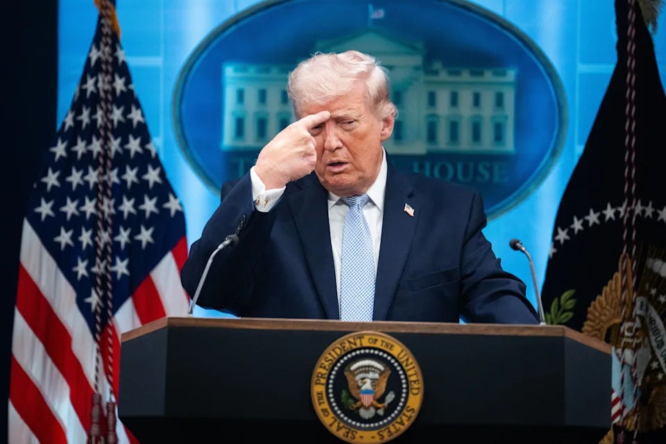 A person at a podium gestures toward their forehead. Two U.S. flags and the presidential seal are visible