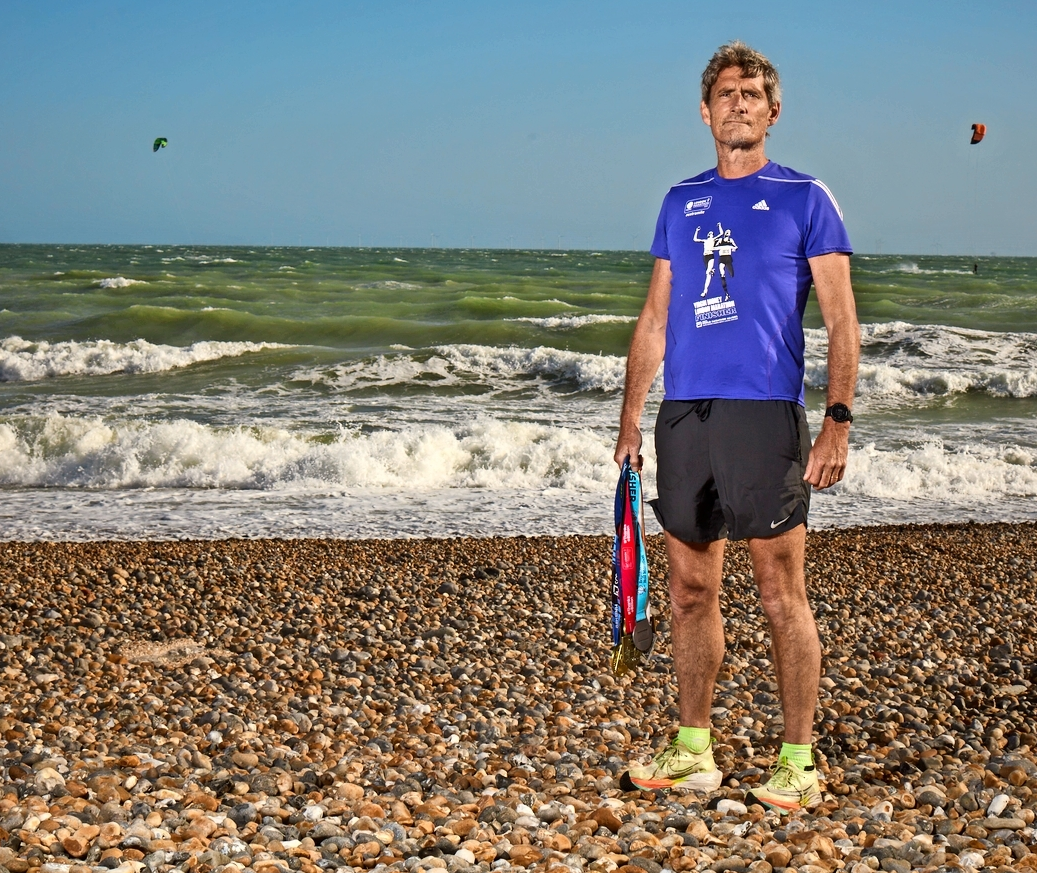 A man in a blue t-shirt and black shorts standing on a pebble beach with crashing waves behind him, holding race medals.