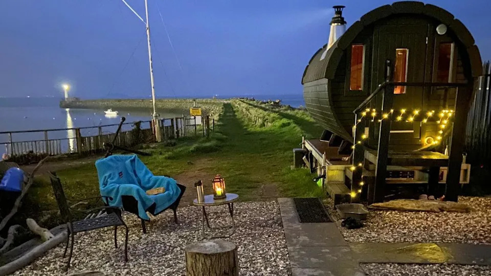 This image shows an outdoor wooden sauna pod in Barry, there's fairy lights wrapped around the sauna. The sauna is based near the beach, you can see the ocean, green grass and dimly lit sky surrounding the sauna. In front of the sauna there's garden chairs and lamps.