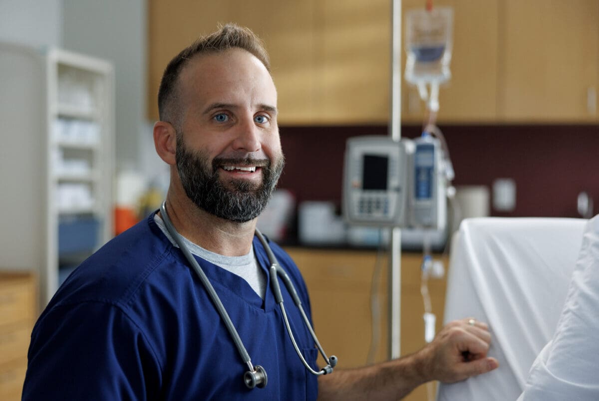 Justin Hicks wears scrubs and poses in front of a patient hospital bed in the nursing SIM lab at UA Little Rock.