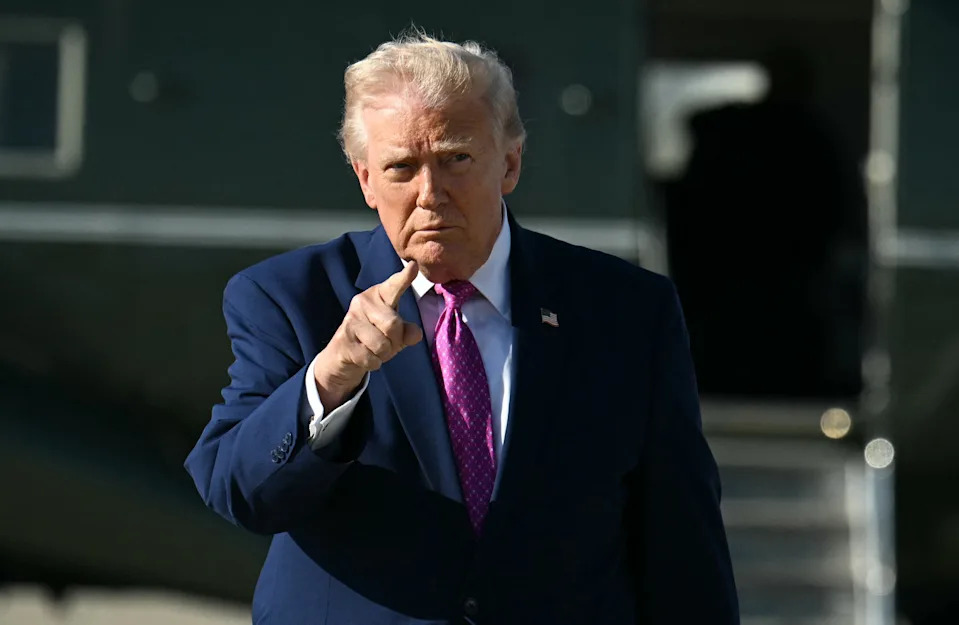 A person in a suit and tie gestures with a finger while walking, with an aircraft in the background