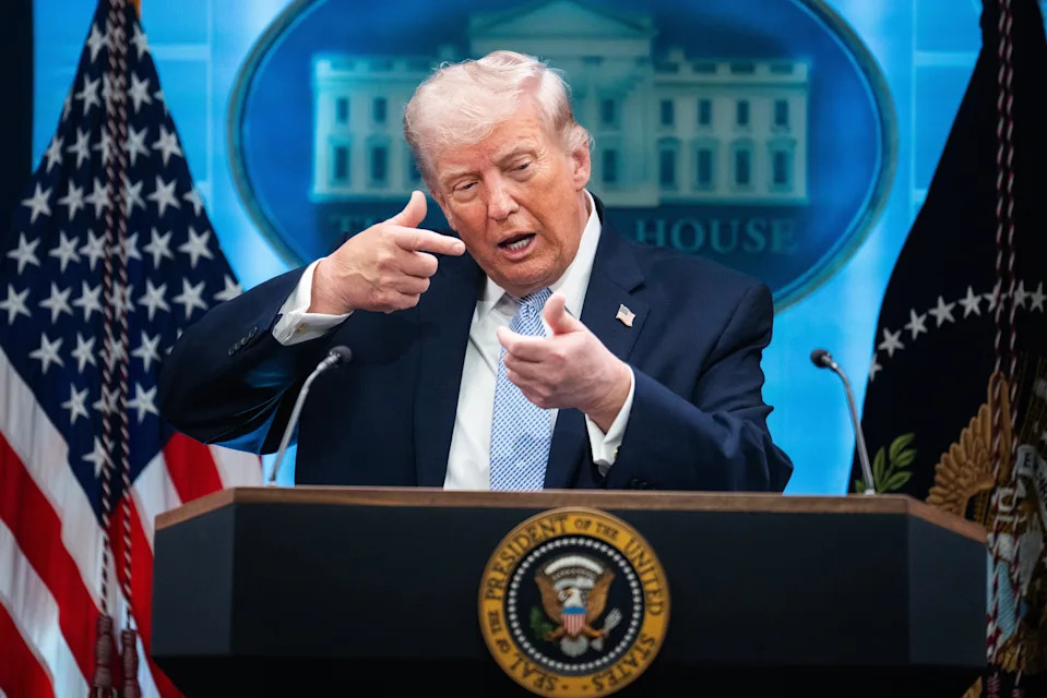 Person at a podium gestures toward the audience with hands shaped like a gun, in front of a White House backdrop