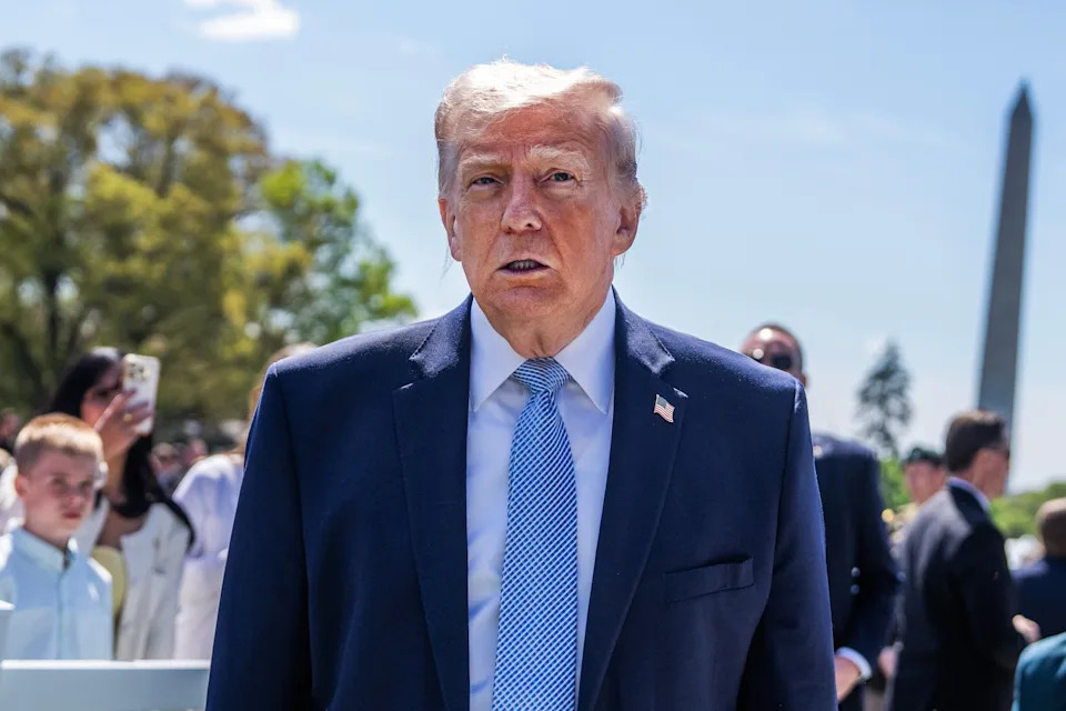 A man in a suit and tie stands outdoors at an event, with the Washington Monument in the background