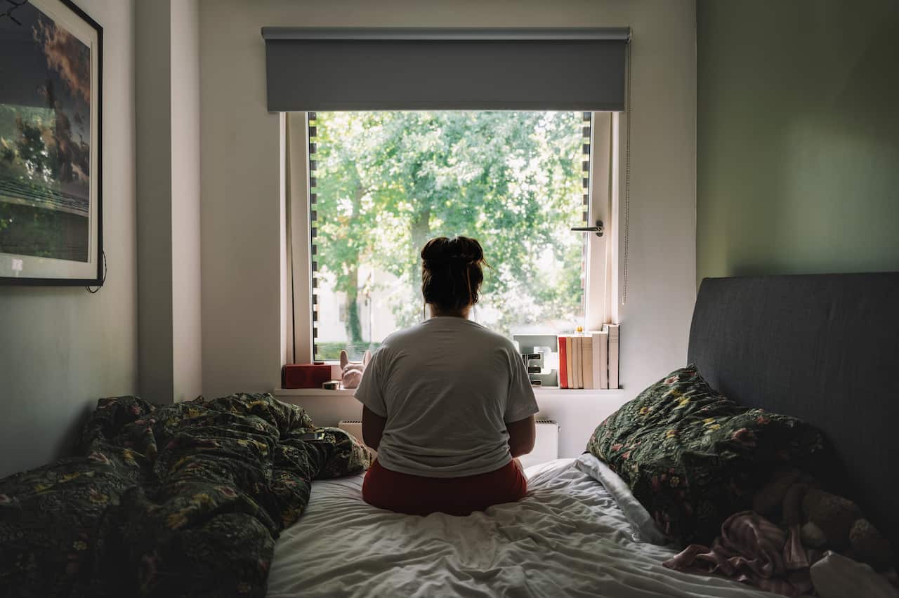 A woman, her back turned, is sitting on her bed, looking out the window.