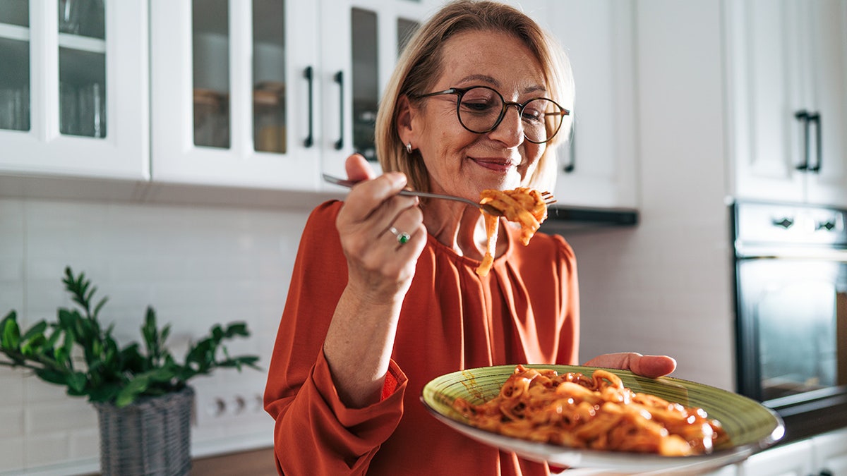 Senior woman smiling and eating pasta twirled on a fork in a kitchen