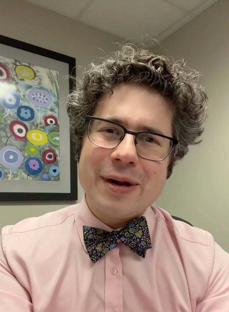 A person with curly gray hair, glasses, a pink shirt, and a colorful bow tie.
