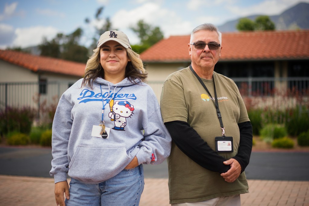 Two people stand side by side outdoors, wearing lanyards, facing the camera.