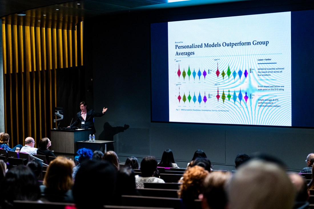 Professor Joshua Curtiss presents his research in front of an audience. On screen says "Personalized Models Outperform Group Averages" with diagrams.