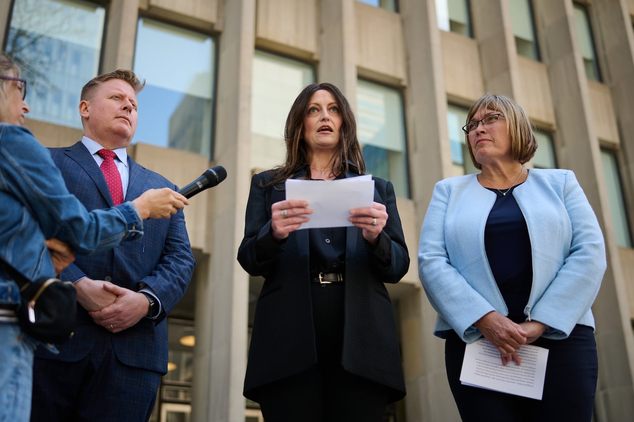 A woman gives a speech outside a courthouse with a man in a suit on her left and a woman in a blue blouse on her right.