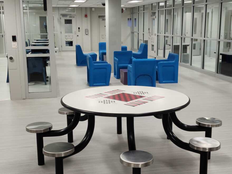 A round table with a chess and checker board painted in the center sits in the middle of an open space inside the men's unit at the St. Charles County Justice Center. Several blue chairs are seen in the background. 