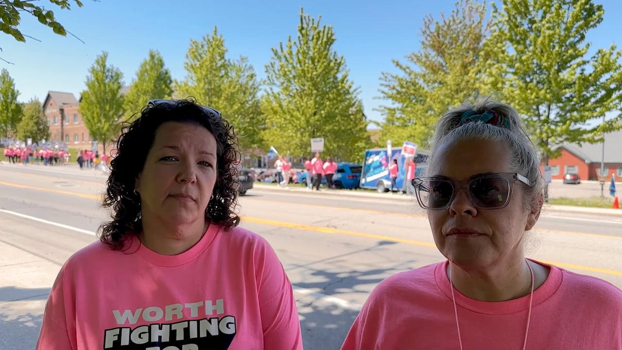 Two women in pink shirts stand across the street from a picket line.