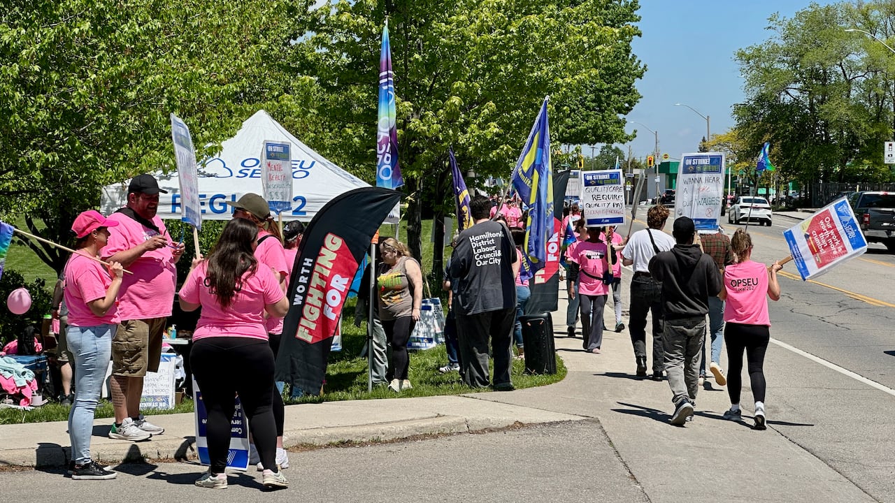 A group of people in pink shirts stand by a tent, many holding picket signs.