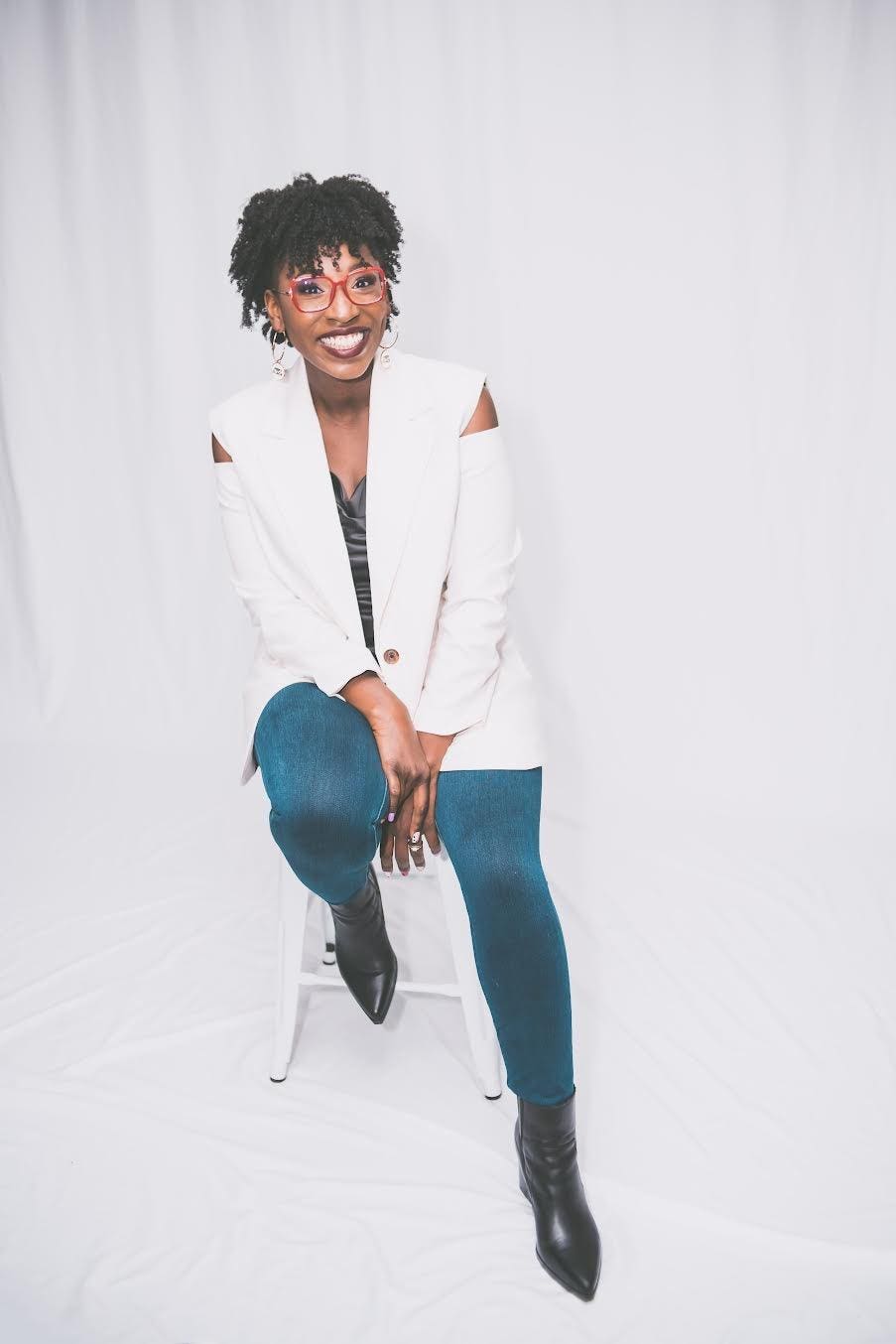Photo of a Black woman smiling with a white blazer, jeans and curly hair with red glasses