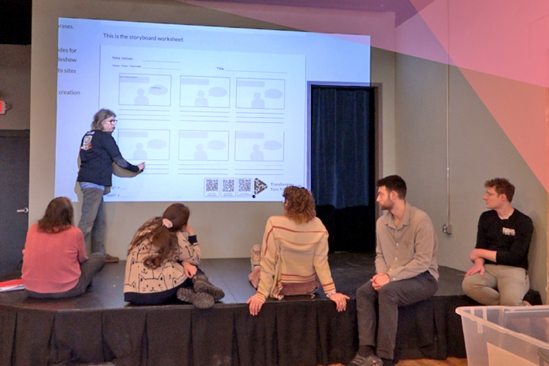 Group of people sitting on a low stage watching an instructor point to a projected storyboard worksheet displayed on a wall in a classroom or workshop setting.