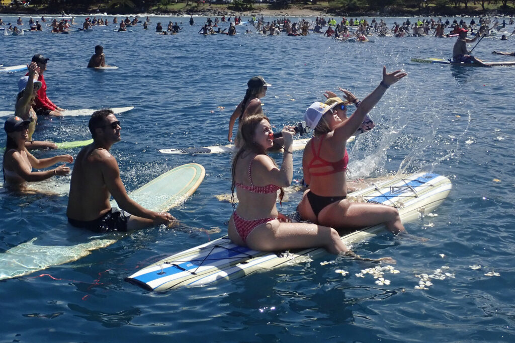 Memorial paddle-out participants from West Maui Kat Lawson films the helicopter dropping flowers while Jennie Hammatt waves off Hanaka‘ō‘ō Beach Park Friday, Aug. 8, 2025, in Lahaina. 102 people died in the Aug. 8, 2023, fire which destroyed the historic West Maui town and Upcountry. (Kevin Fujii/Civil Beat/2025)