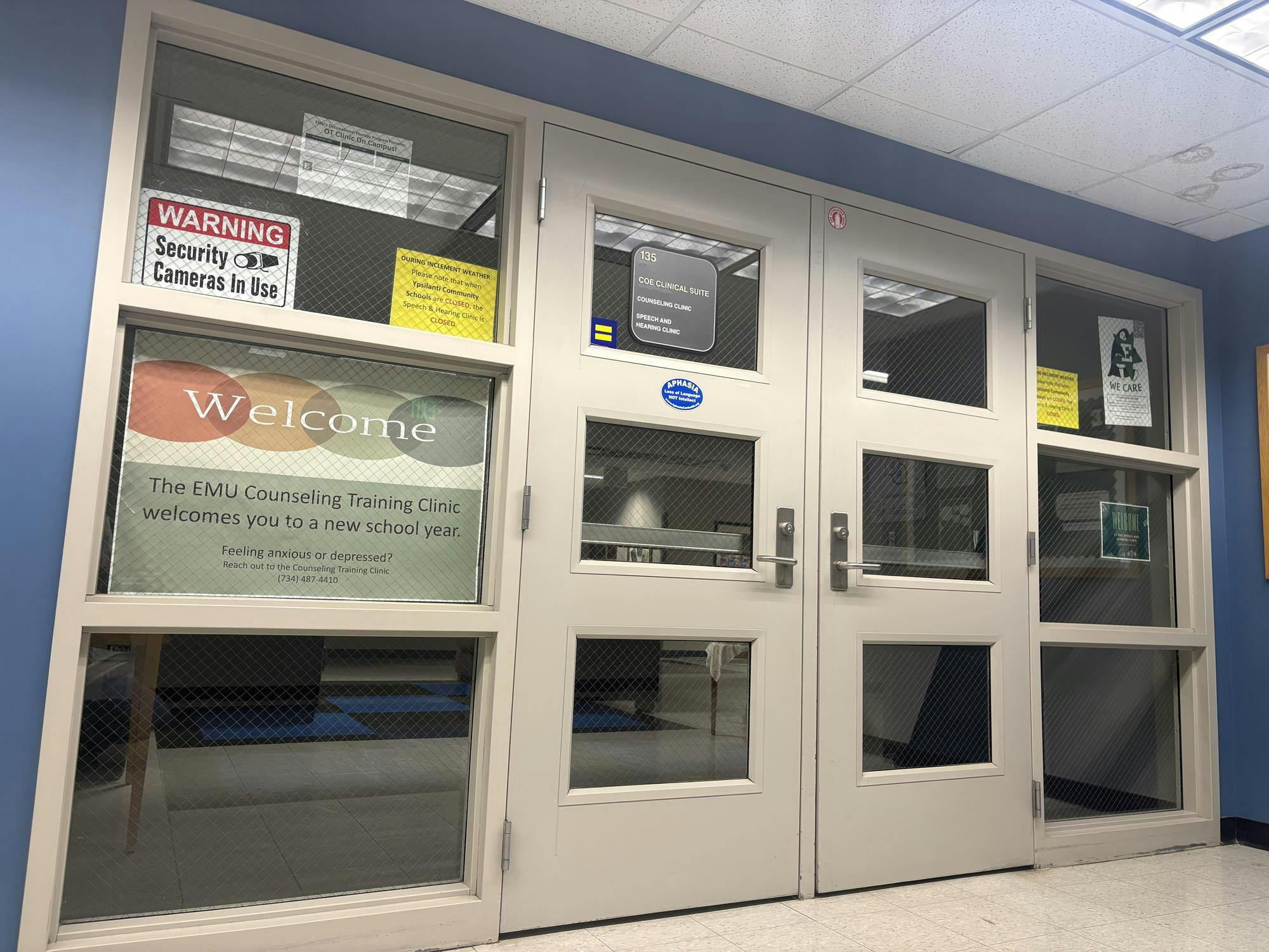 Two white closed doors in a blue wall. Windows around the doors have various posters inside.