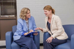 Two women in business attire sit side by side in blue chairs and talk, with one holding a clipboard and pen.