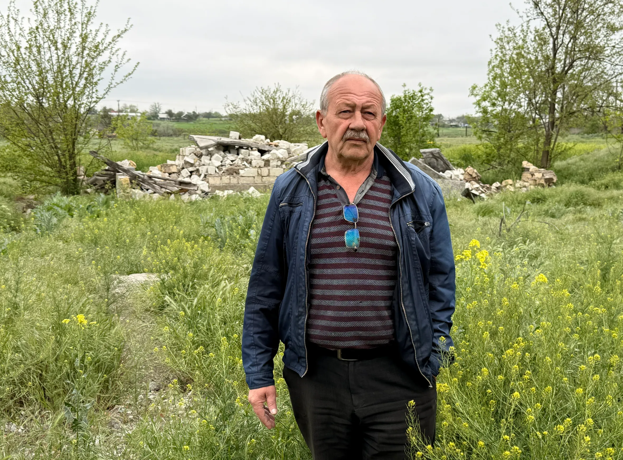 Ivan Volko, the village elder, stands in a field of yellow flowers and green plants, with rubble in the background.