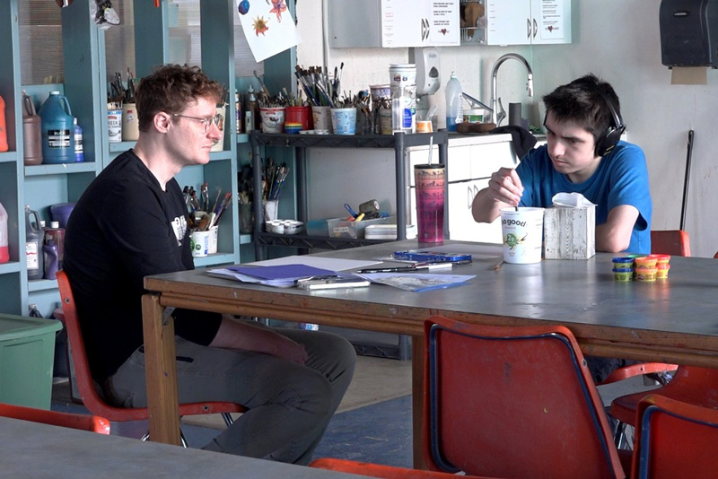 Two individuals seated at a large table in an art classroom, surrounded by paint bottles, brushes, and supplies, while one stirs material in a container and the other looks on.