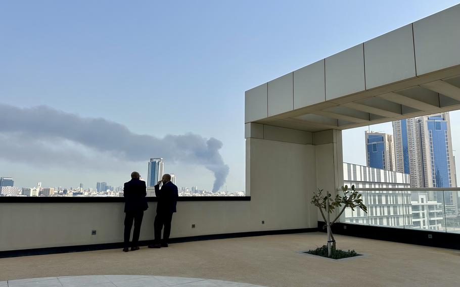 Two people look out from a rooftop terrace toward a rising smoke plume.