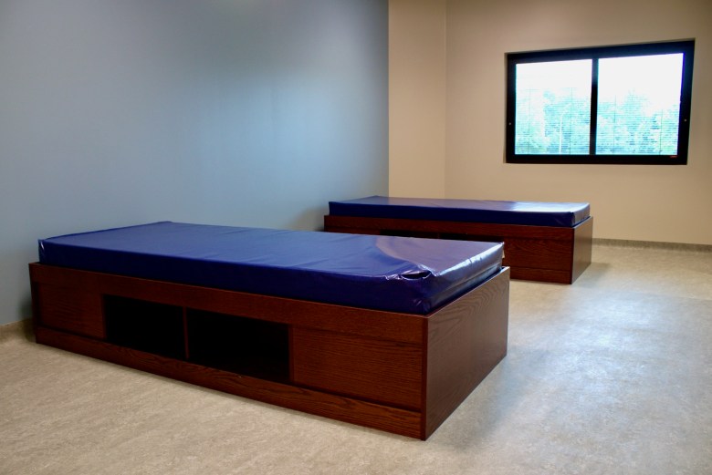 A window casts light onto two blue twin XL mattresses that sit on wooden bedframes in an inpatient psychiatric unit.