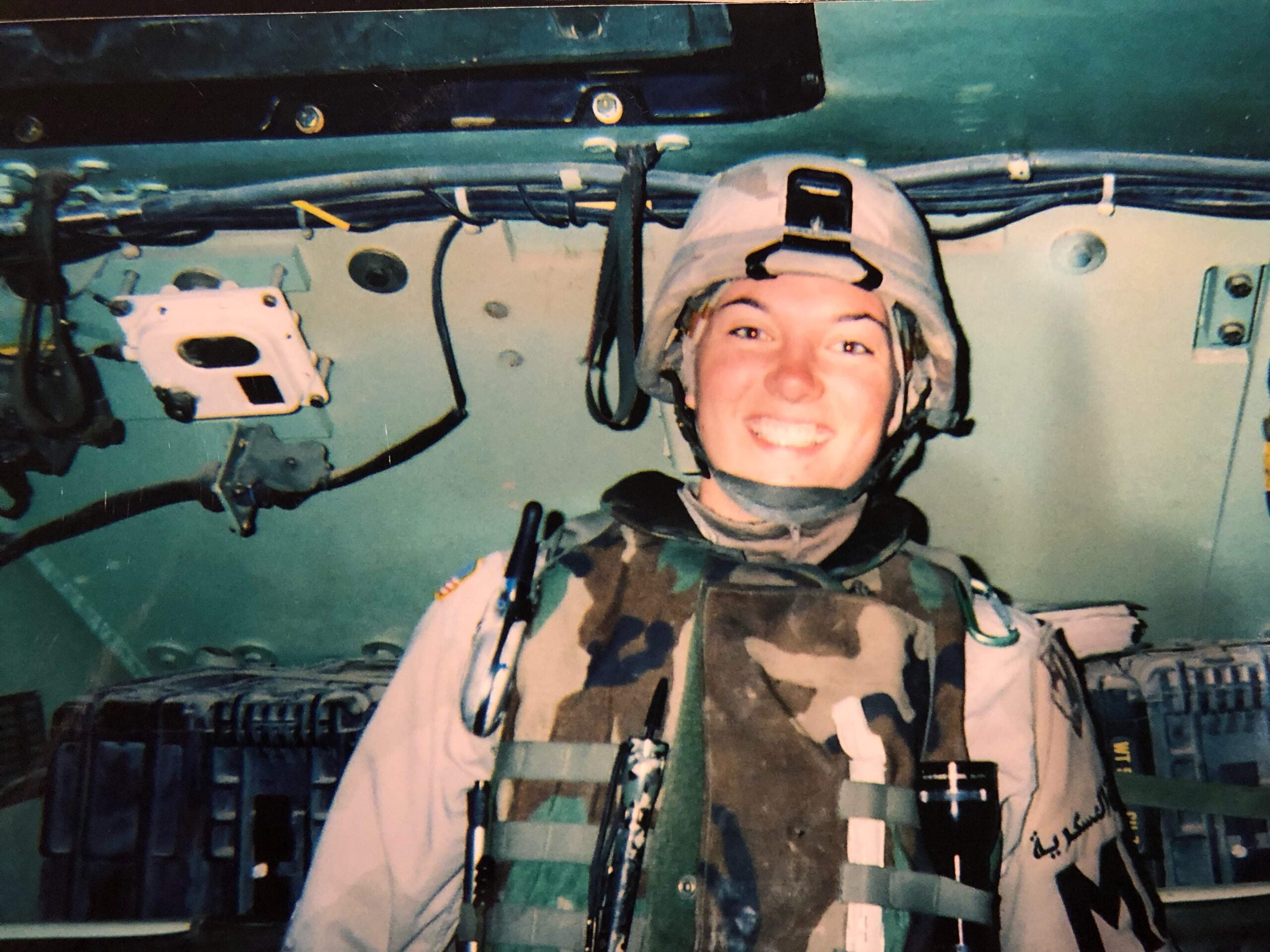 A person in military uniform and helmet smiles inside an armored vehicle, surrounded by cables and equipment.