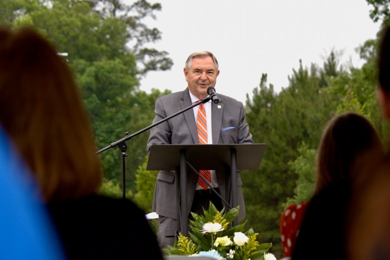 A white man with gray hair and wearing a gray suit and an orange tie speaks into a microphone in front of a podium.