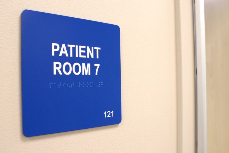 A blue sign reads “Patient Room 7” on a cream wall inside a psychiatric facility.