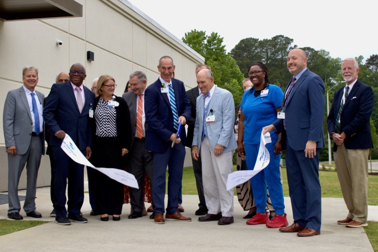 A group of men in suits and a woman in blue scrubs cut a ceremonial ribbon with large scissors outside a health care facility.
