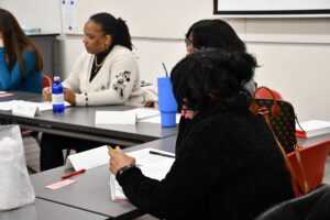 A Black woman with a ponytail talks to a group at a table. 