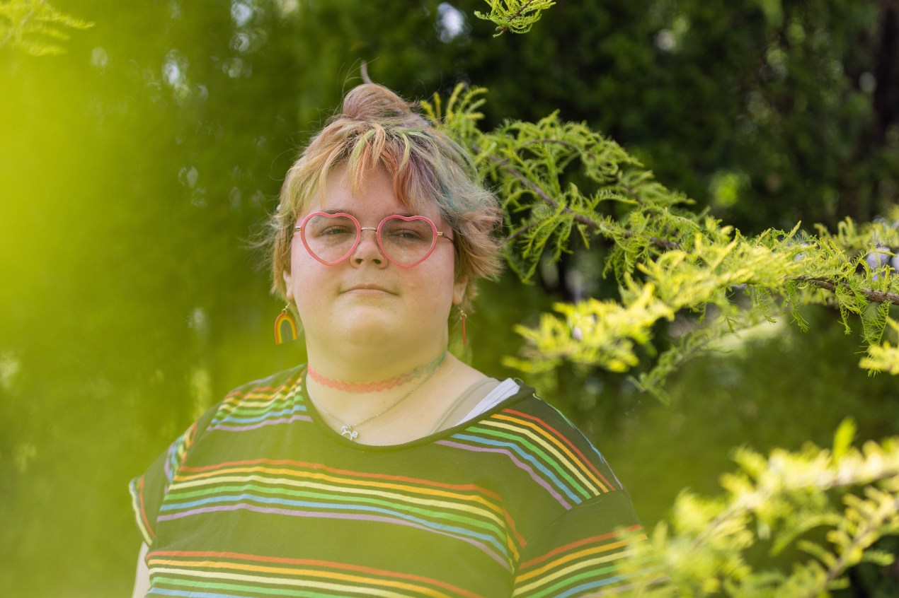 A portrait of a young person wearing a rainbow T-shirt, rainbow earrings, and heart-shaped glasses. Green foliage frames the photograph.