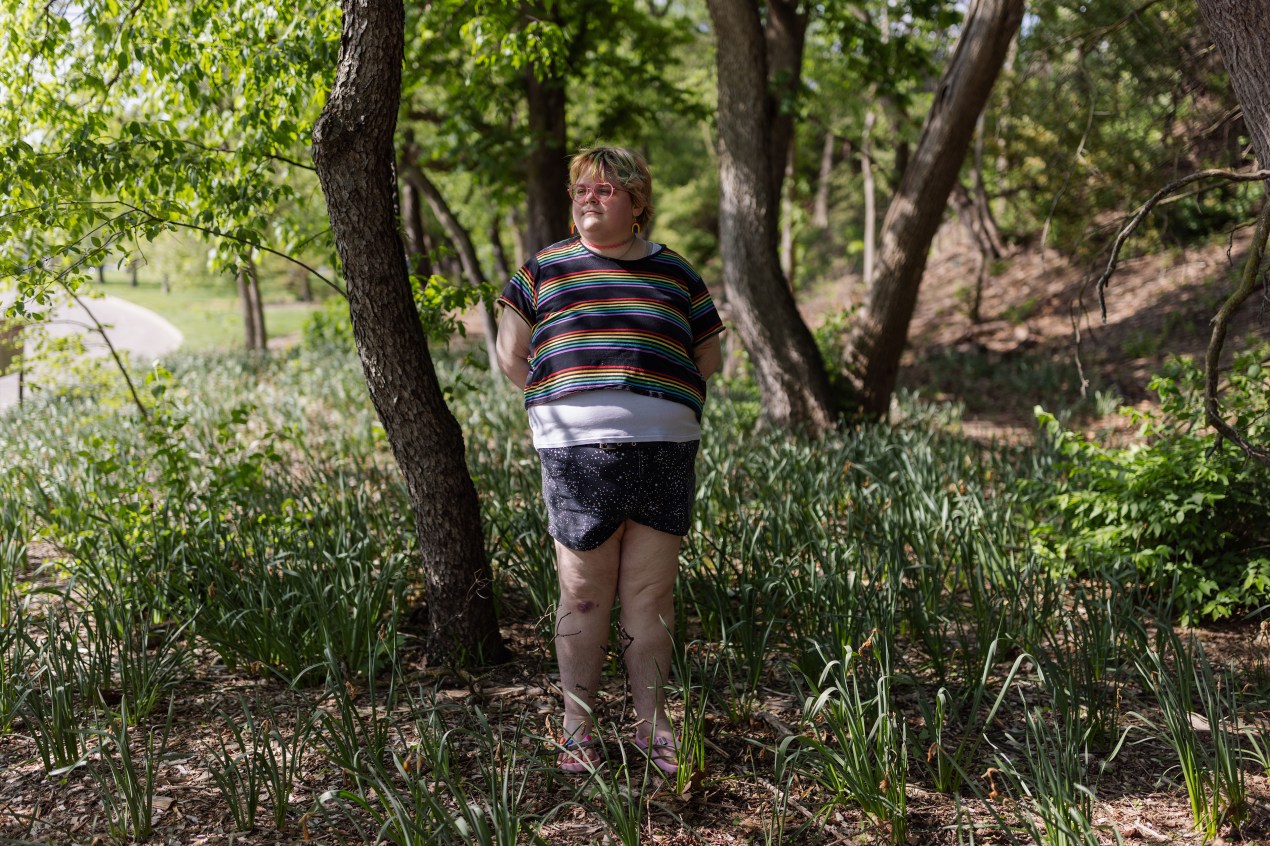 A young person wearing a rainbow T-short, shorts, and heart-shaped glasses stands amongst trees and tall green grasses.