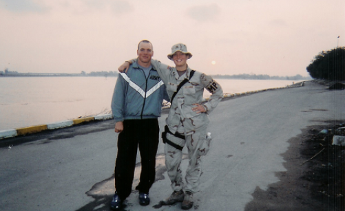 Two people in military attire stand side by side on a paved road near a body of water, with one person’s arm around the other. The sky appears overcast.
