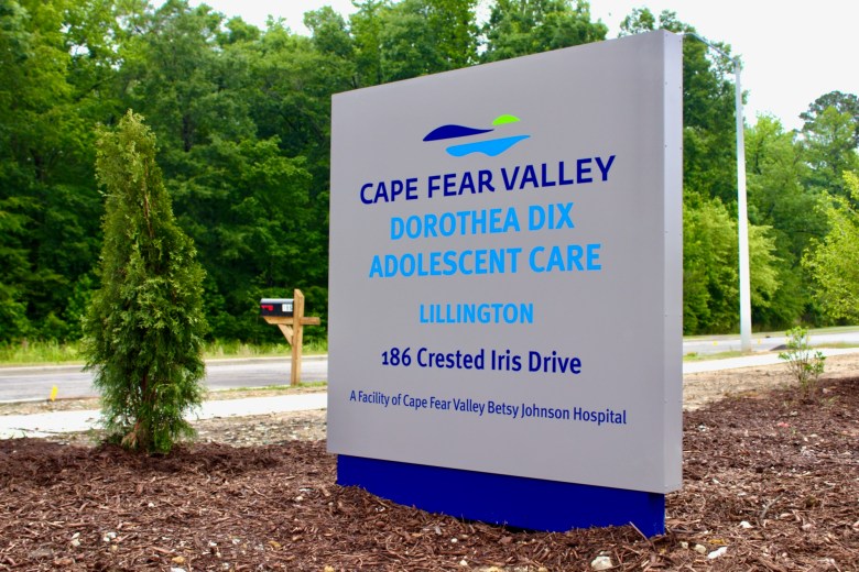 A gray sign sitting in mulch reads “Cape Fear Valley Dorothea Dix Adolescent Care – Lillington 186 Crested Iris Drive A Facility of Cape Fear Valley Betsy Johnson Hospital.”