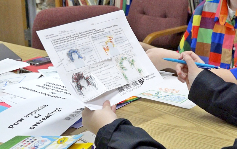 Person holding a worksheet with hand-drawn images and notes at a table covered with papers and art supplies, wearing a brightly colored patterned shirt and holding a blue pen.