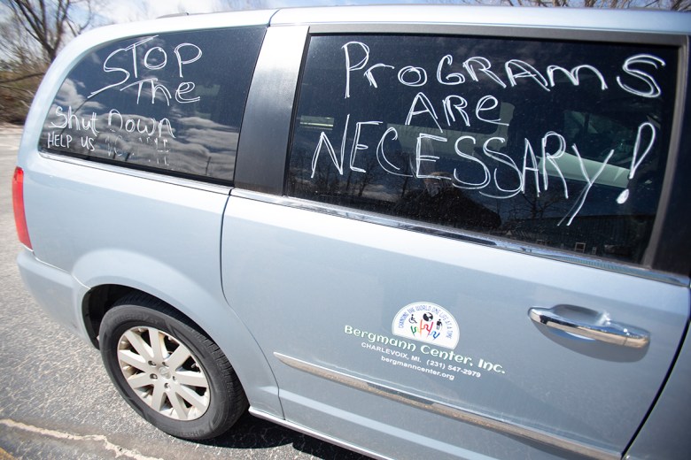 A van from Bergmann Center Inc. is parked outdoors with messages written on the windows reading “STOP the shutdown” and “Programs are necessary!” in white marker.