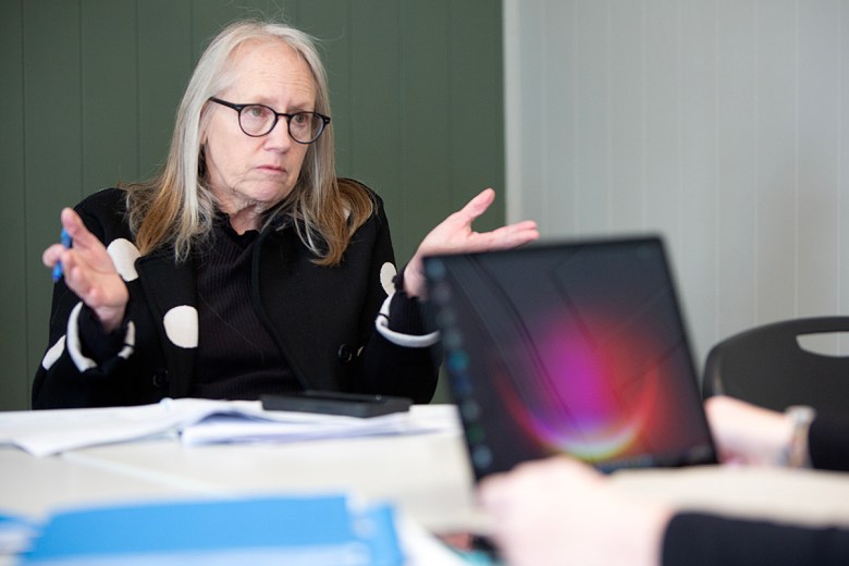 A woman wearing glasses gestures while speaking during a meeting, seated at a table with papers and a laptop in the foreground.