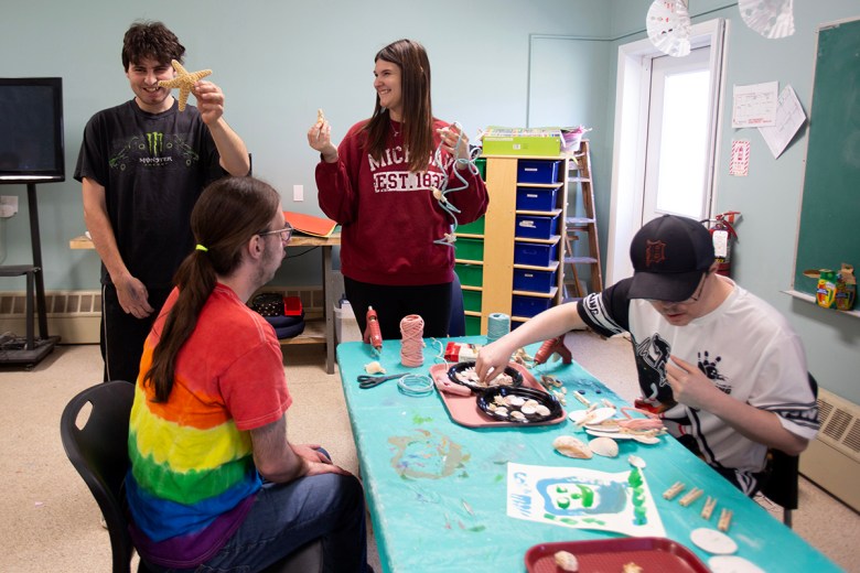 Four people gather around a craft table in an activity room, smiling and working with seashells, string and art supplies while one person holds up a starfish decoration.