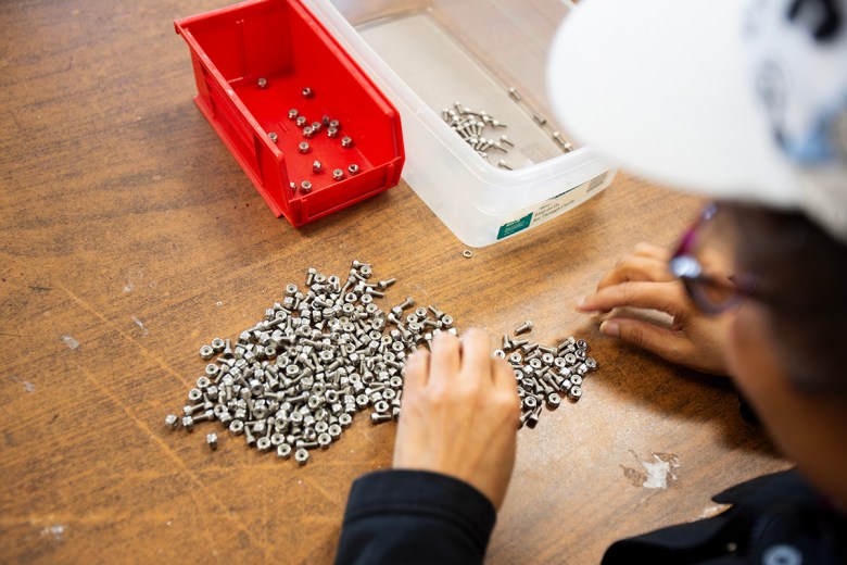 A person sorts small metal nuts and bolts into plastic bins on a wooden table during a hands-on  activity.