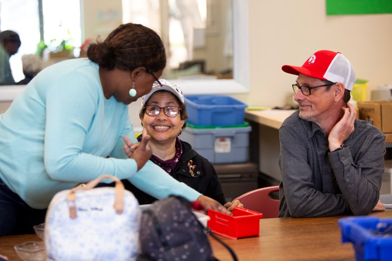 Three people smile and talk around a table in a community workspace, with plastic bins and supplies spread across the room.