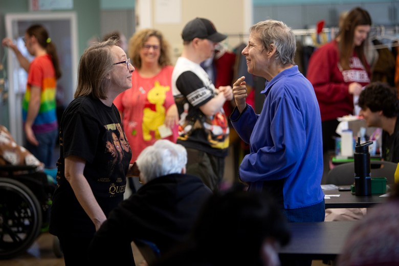 Two women smile and talk inside a busy community activity room, while other people socialize and work at tables in the background.