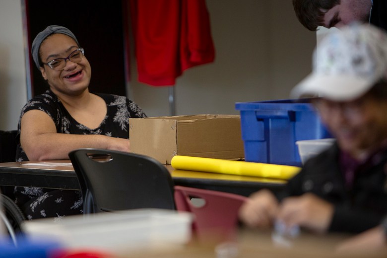 A smiling woman wearing glasses and a headband sits at a table in a classroom or activity space, with boxes and bins nearby as other people work in the blurred foreground and background.