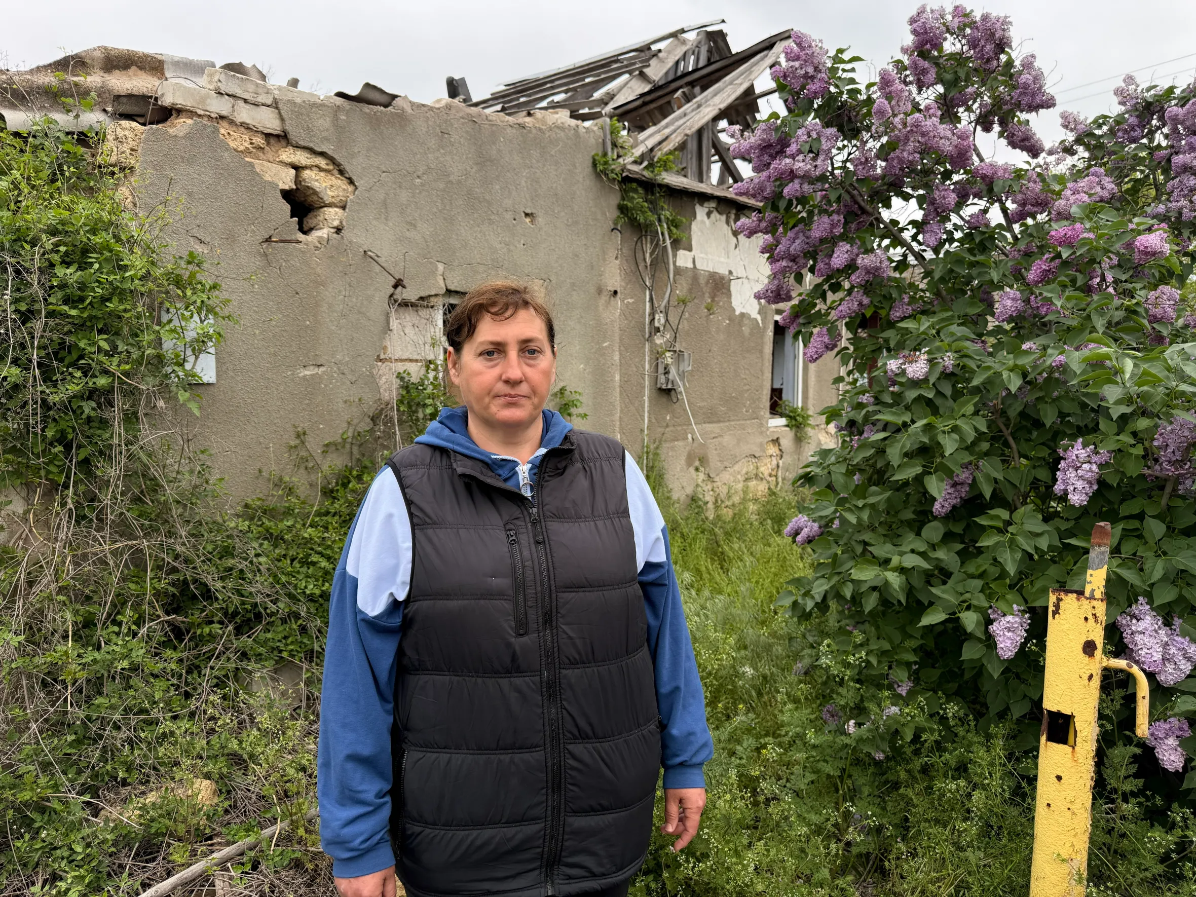 Olha, a local, stands in front of a partially destroyed building and a lilac bush.
