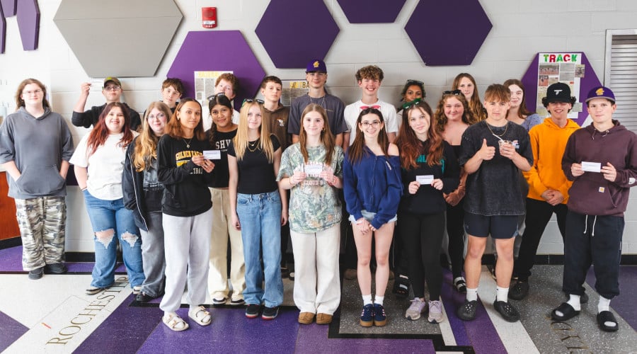 Over two dozen students pose in a school hallway, many holding small white cards.