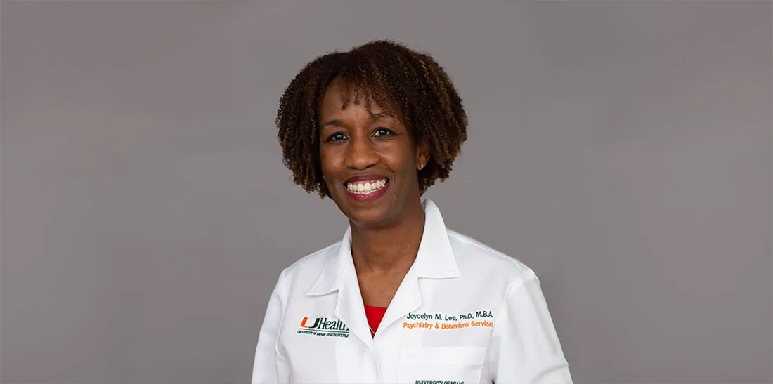 Portrait of Dr. Jocelyn Lee wearing a white UHealth-branded lab coat, standing against a plain gray background. The lab coat features embroidered text on the left chest that reads “Jocelyn M. Lee, PhD, MBA” and “Psychiatry & Behavioral Sciences,” along with the University of Miami Health System (“UHealth”) logo on the opposite side. The person is facing forward with visible teeth in a smile, and has short, tightly curled hair. The image is evenly lit with no visible shadows or background elements.
