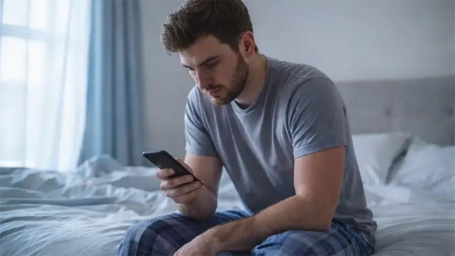 An unrested man sitting on the edge of a bed, looking stressed while checking his phone shortly after waking up.