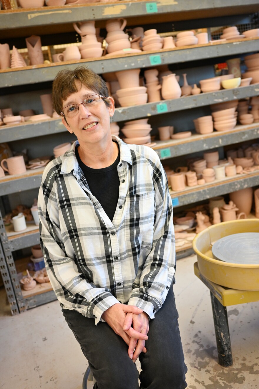 A woman is seated in front of shelves of pottery.